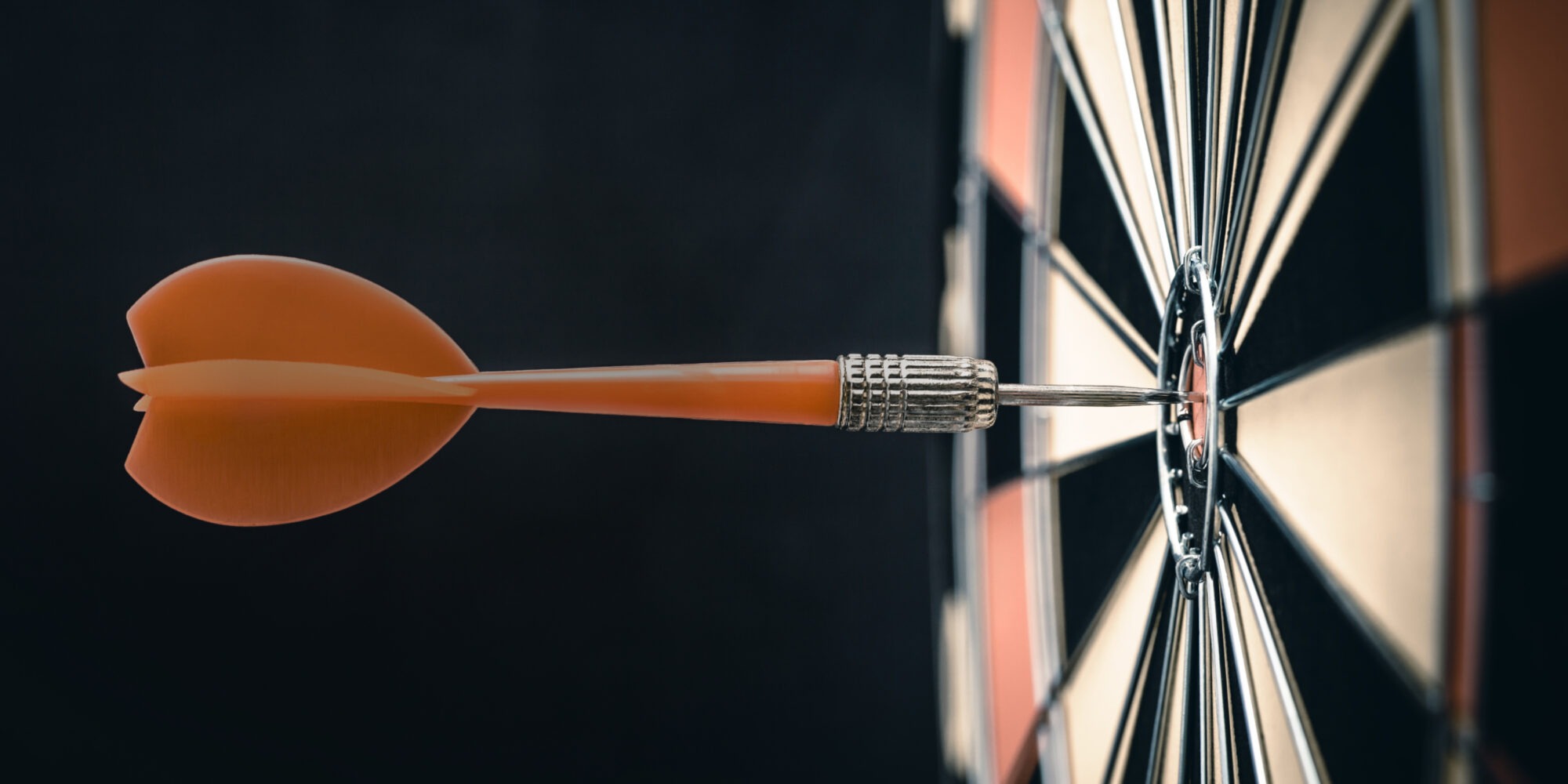 Nahaufnahme eines Dartpfeils, der exakt die Mitte einer Dartscheibe trifft (Bullseye), vor dunklem Hintergrund